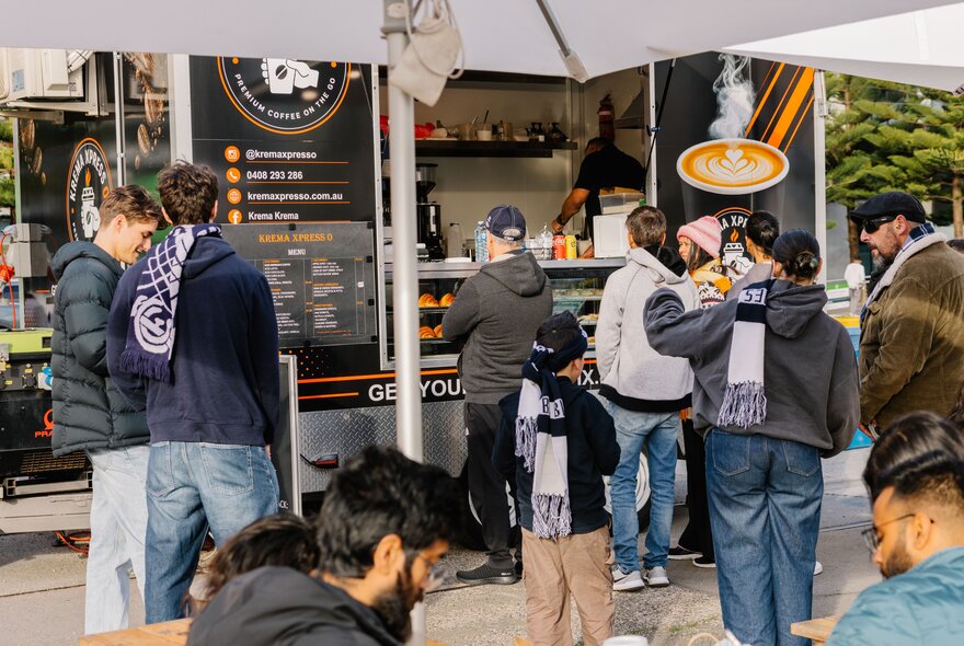 People waiting in line at an outdoor food truck, with several individuals wearing matching AFL football club scarves.