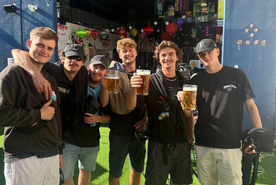 A group of six young men holding up large beers in a bar setting.