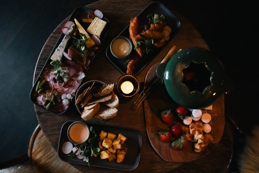 Overhead view of a table with a cheese platter, a chocolate fondue and platers of cut up fruit, bread and drinks.