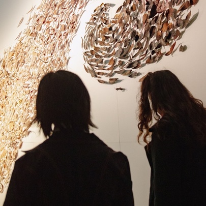 The silhouettes of two people looking at an Indigenous art installation of gum leaves on the wall.
