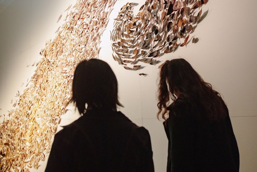 The silhouettes of two people looking at an Indigenous art installation of gum leaves on the wall.