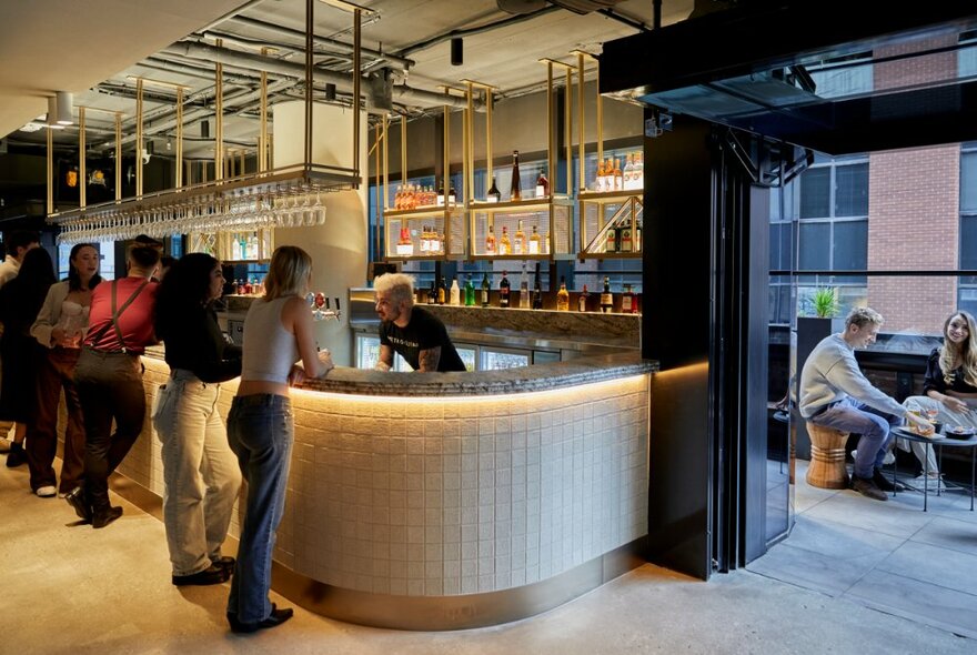 People standing at an illuminated bar in a pub setting, looking on to balcony with outside seating.