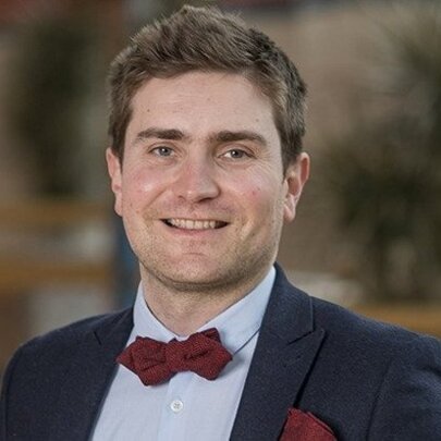 A smiling man with brown hair, identified as Thomas Roulet, dressed in a navy blazer over a light blue shirt and a red bow tie.