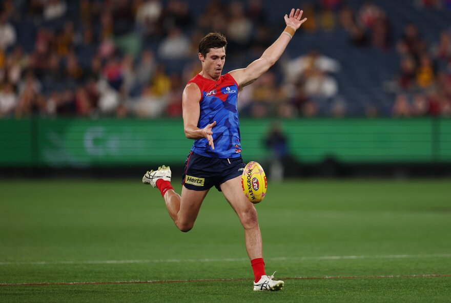 A Melbourne AFL player running while about to kick a yellow football he has just dropped in front of his leg.
