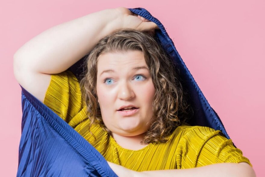 A woman stretching a blue pleated skirt over her head while wearing a mustard-coloured top.