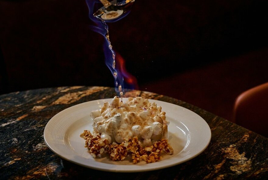 Dessert being lit by small blow torch on dark marble-look surface.