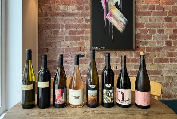 Bottles of wine lined up in a row on a restaurant dining table, an exposed brick wall in the background.