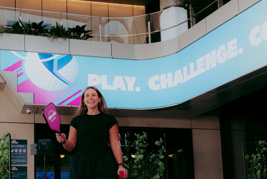 A person smiling and holding a pink pickleball paddle inside Marvel Stadium.