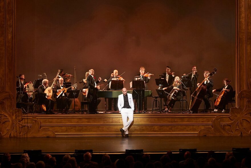 A singer wearing a light coloured suit standing casually in front of a performing orchestra on stage.