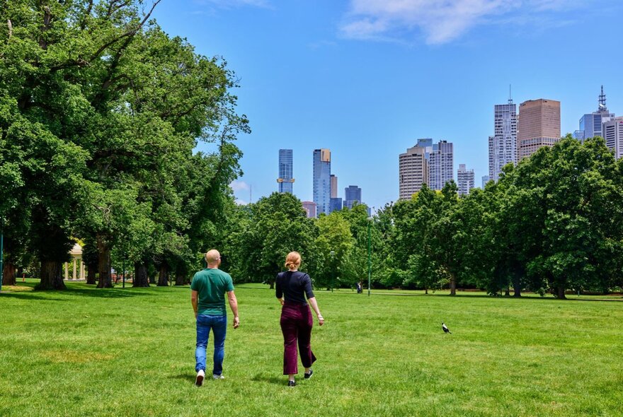 People walking through a grassy park, with city buildings on the blue sky horizon. 