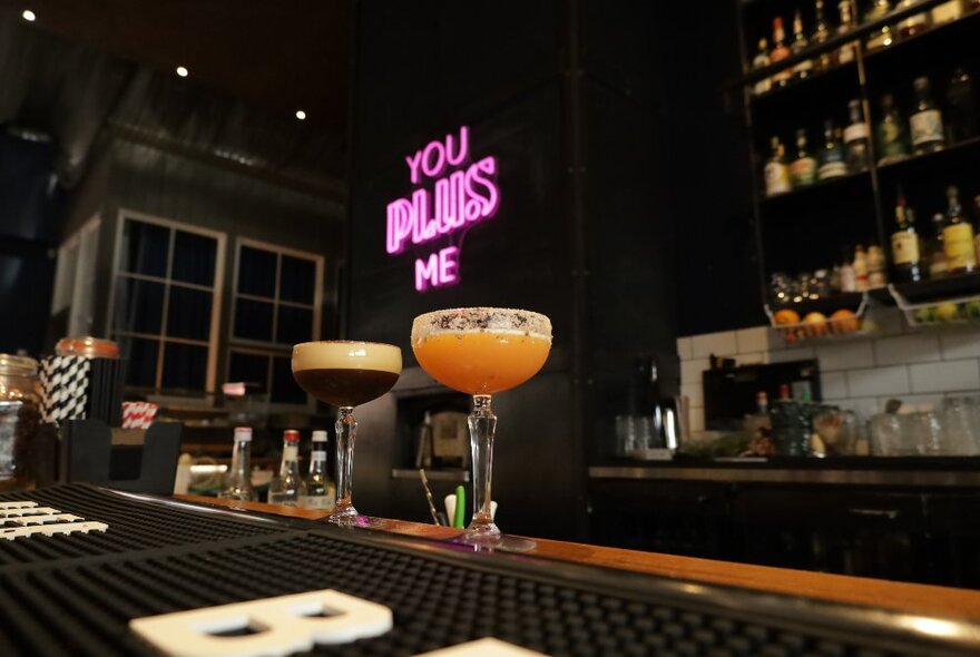 Cocktails on a bar counter with pink neon sign and shelves of bottles in the background.