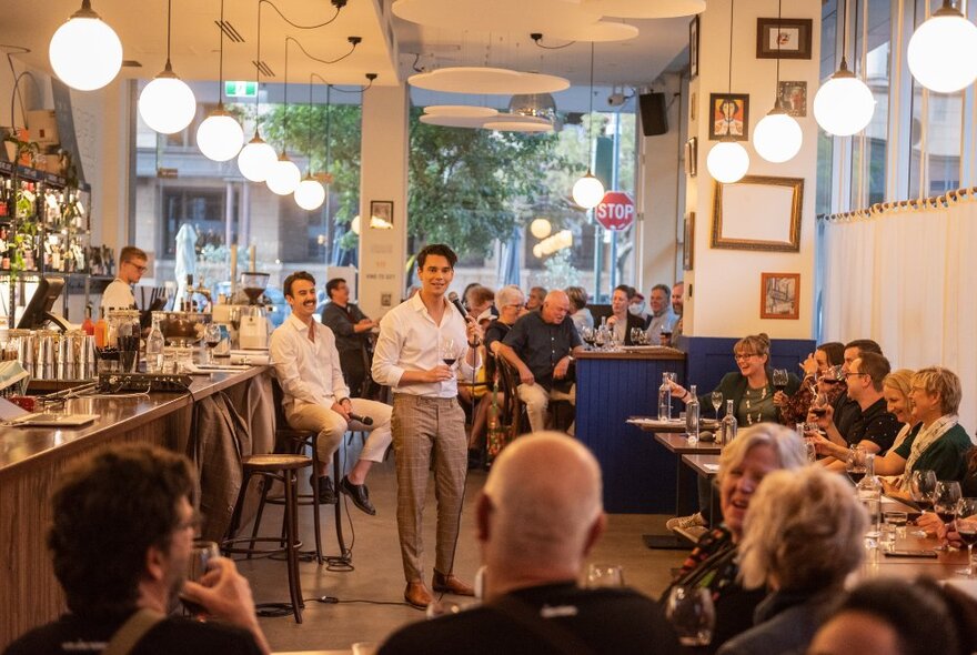 A man walking and talking in front of a seated audience inside a bar.