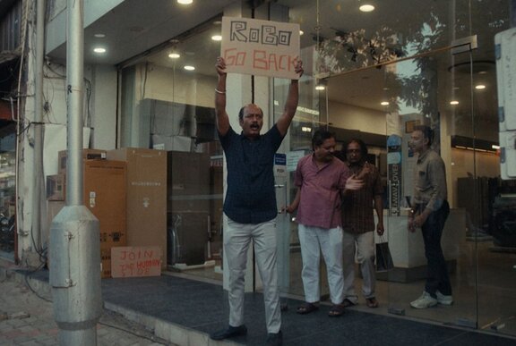 Screen still from an Indian film: a man holds a banner overhead outside a shop.