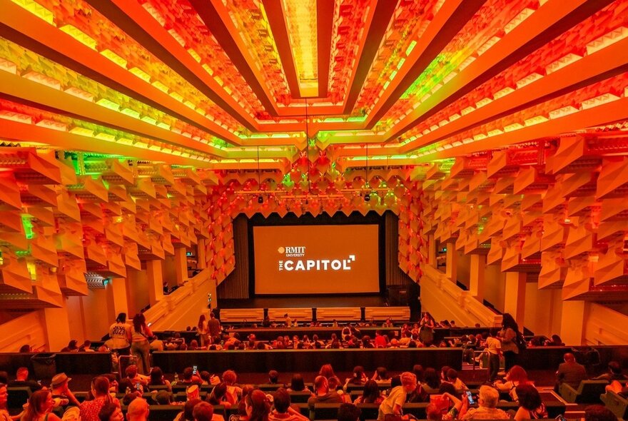 Capitol Cinema with red and orange lighting, seated audience in front of large screen with signage.