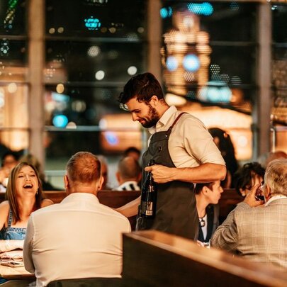 A waiter attends a table with a woman laughing in a restaurant at night. 