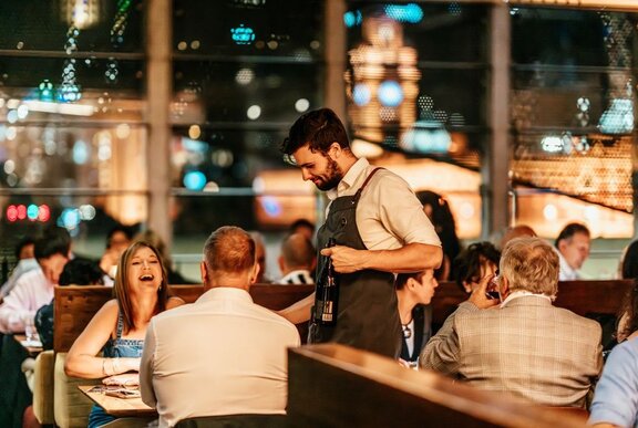 A waiter attends a table with a woman laughing in a restaurant at night. 