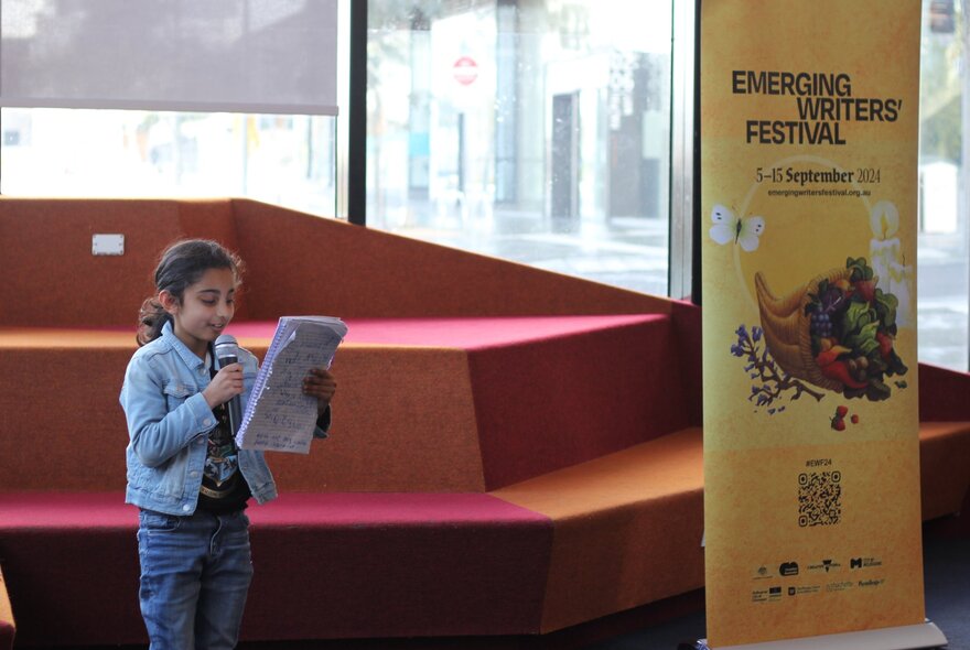 A school aged child holding a microphone and reading from a notepad in a venue, a banner to the right of her is a sign for the Emerging Writers' Festival.