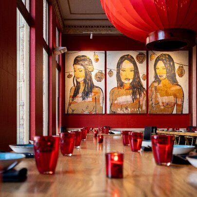 Interior of Red Spice Road with long wooden table and chairs, red ceiling lantern and three portraits at back of room.