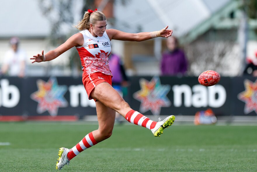 A Sydney Swans AFLW player kicking a football on the field, with her arms outstretched and kicking leg raised off the ground.