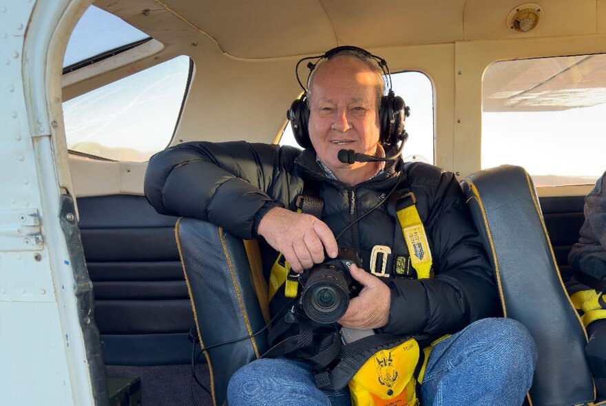 A photographer holding a camera in a seat of a helicopter with a headset and mic on. 