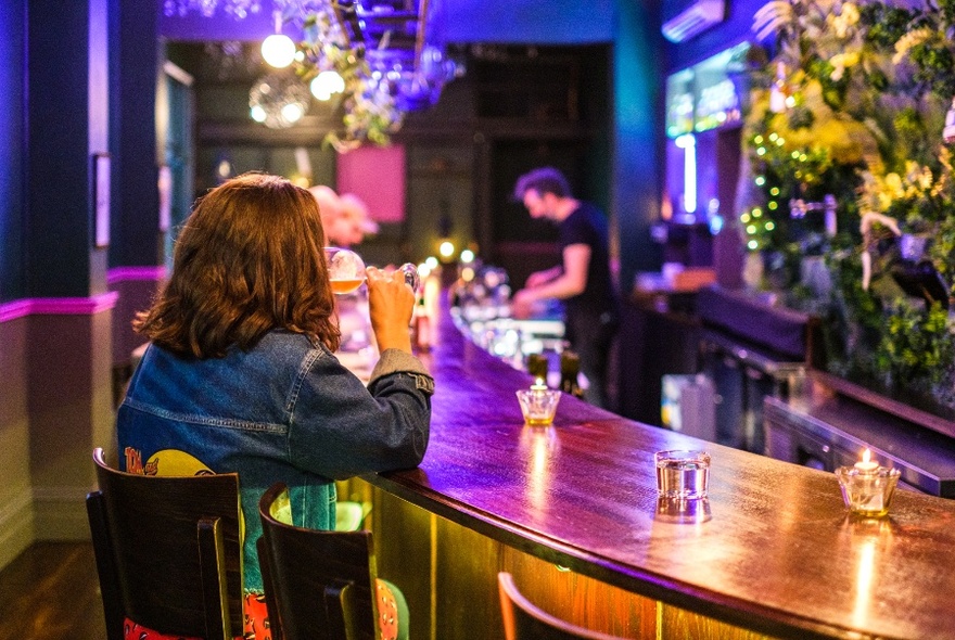 Curved wooden bar, decorated with small tealight candles, with long-haired patron drinking, other patrons and a bartender serving drinks at rear.