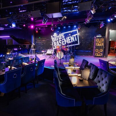 Intimate basement jazz club featuring a brightly lit stage with a neon sign that reads 'Bird's Basement'. The venue is filled with elegant blue velvet dining booths and tables set under dim, atmospheric purple lighting.