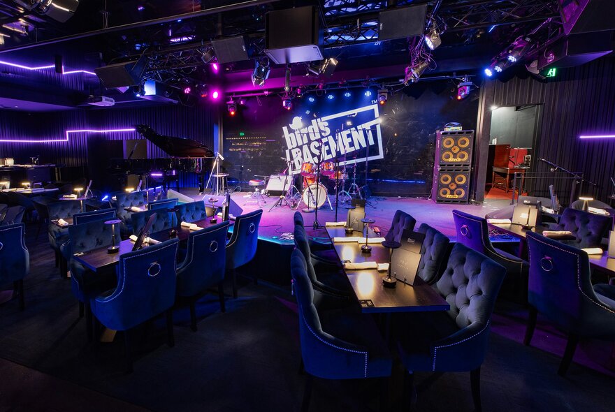 Intimate basement jazz club featuring a brightly lit stage with a neon sign that reads 'Bird's Basement'. The venue is filled with elegant blue velvet dining booths and tables set under dim, atmospheric purple lighting.