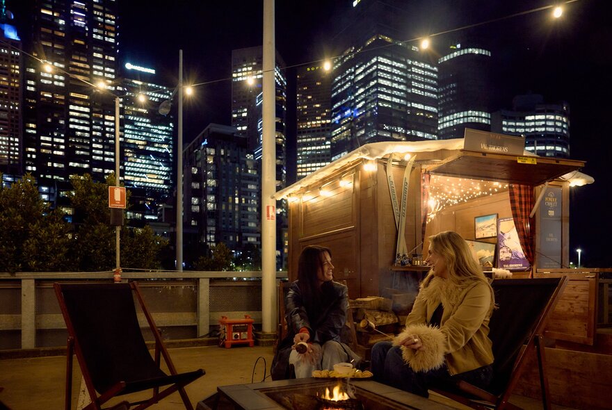 Two friends are sitting outside a small wooden chalet. There is a firepit and fairy lights and a view of the city skyline behind them.
