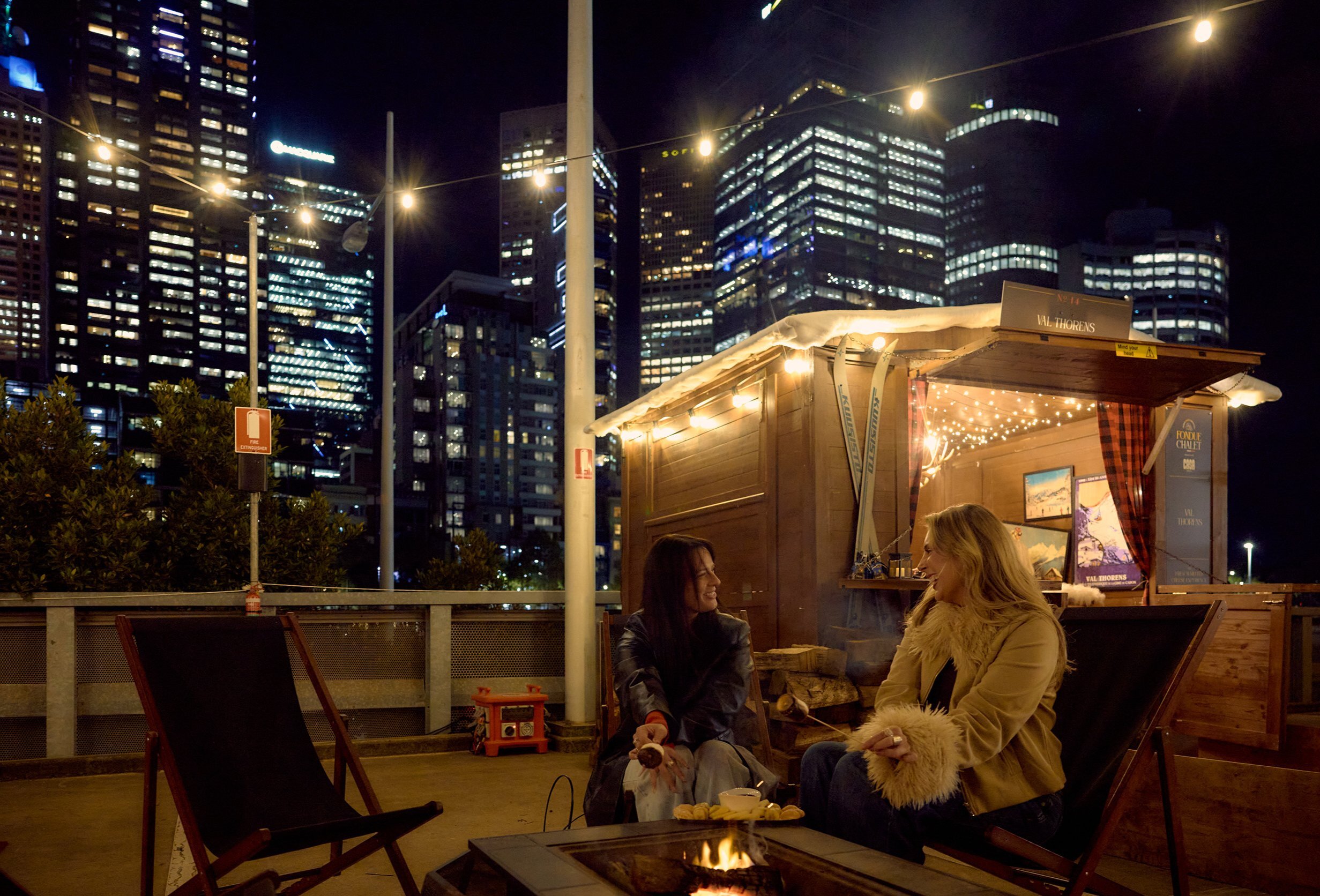 Two friends are sitting outside a small wooden chalet. There is a firepit and fairy lights and a view of the city skyline behind them.