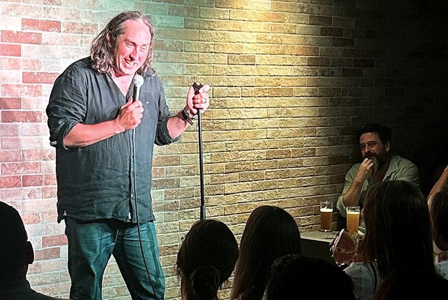 Comedian Ross Noble performing stand-up at a small venue with a brick wall background. 