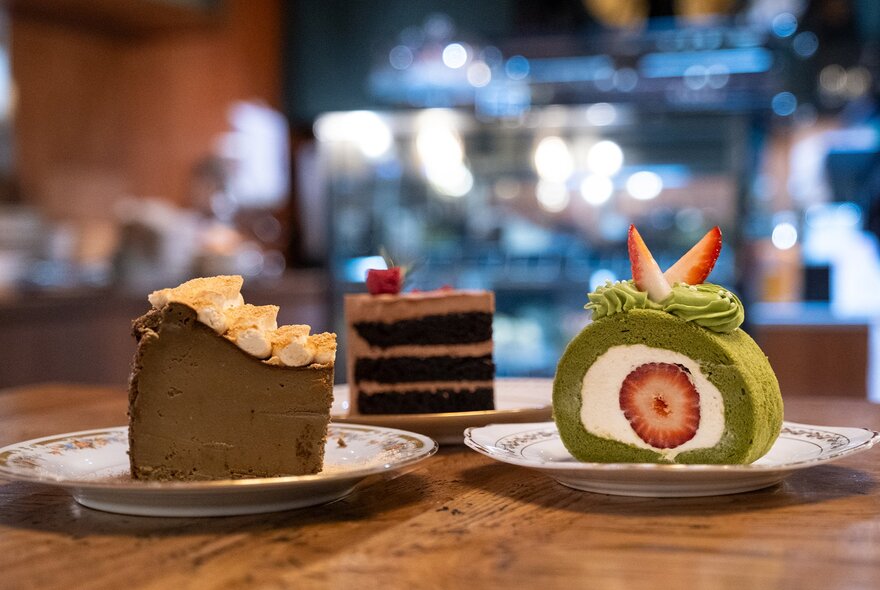 Three cake slices on a table including a brown cheesecake, matcha and strawberry swiss roll and a layered chocolate cake.