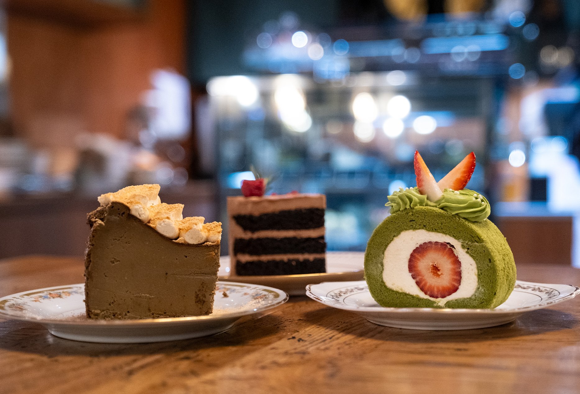 Three cake slices on a table including a brown cheesecake, matcha and strawberry swiss roll and a layered chocolate cake.