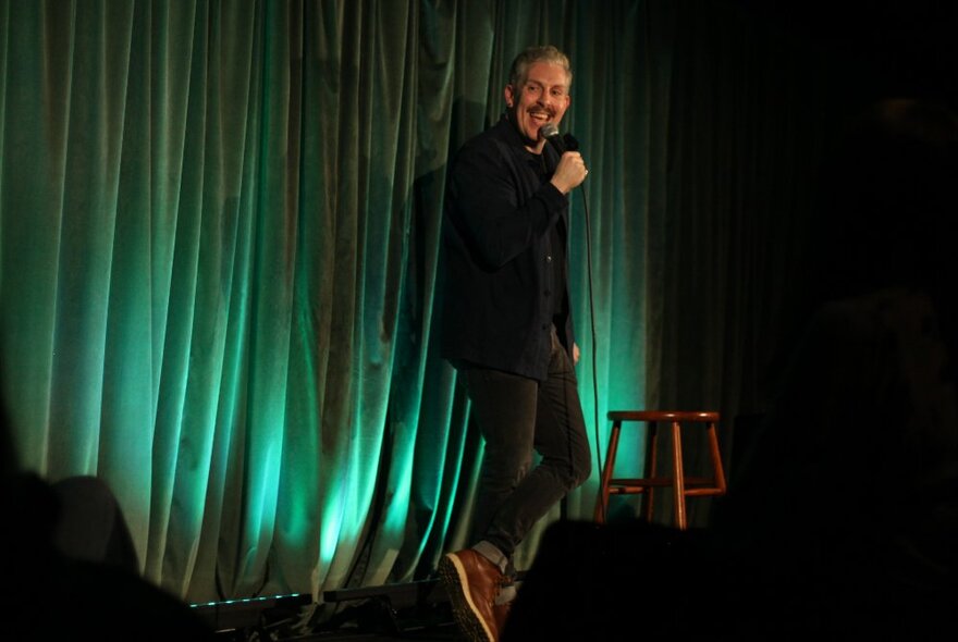 A male comedian standing on a stage in front of an audience, holding a microphone and performing stand-up comedy.