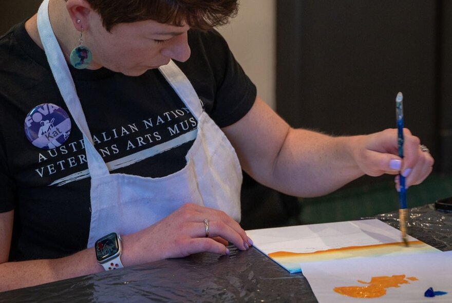 A person wearing an apron, seated at a table as part of am art workshop, using a paintbrush to apply paint to paper.