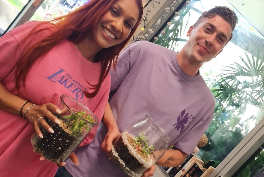 Two people holding glass-domed terrariums, shot on an angle with plants in the background.