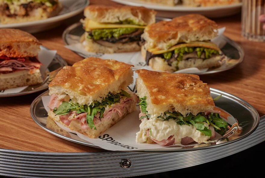 Filled focaccia sandwiches on silver trays, on a wooden table in a cafe.