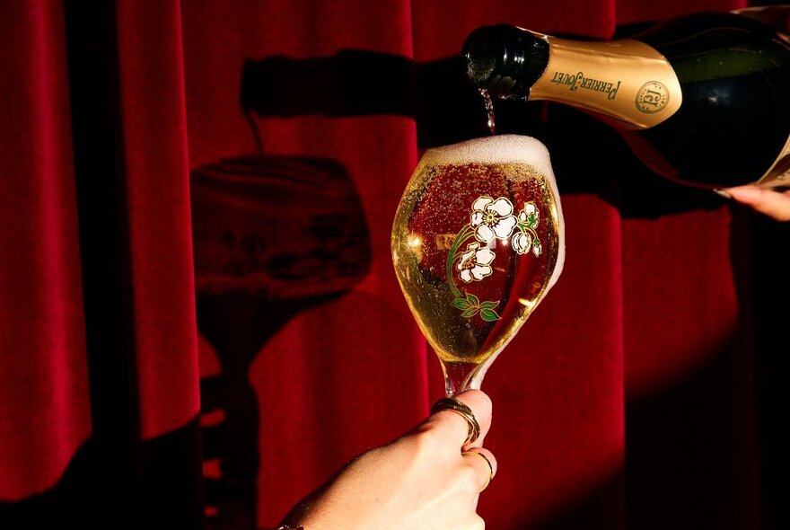 A glass being filled to the brim with French Champagne, against a red velvet curtain.