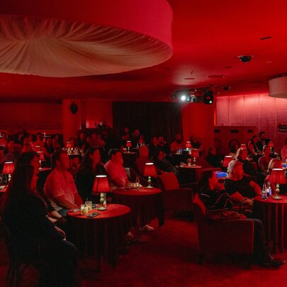 A group of people seated in a small, intimate venue with red lighting and small red lamps on tables. 