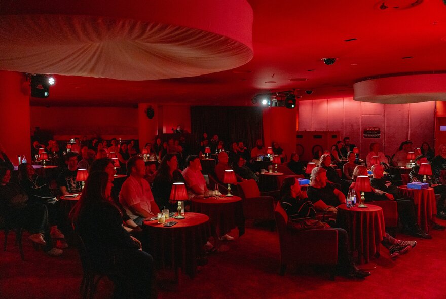 A group of people seated in a small, intimate venue with red lighting and small red lamps on tables. 
