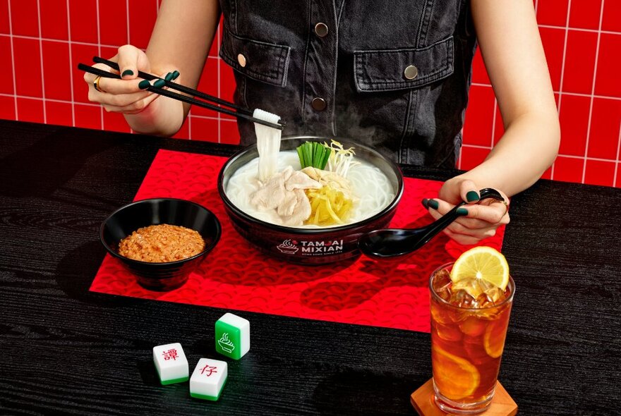 Looking down on a table with a person lifting noodles from a bowl with condiments and ice tea. 