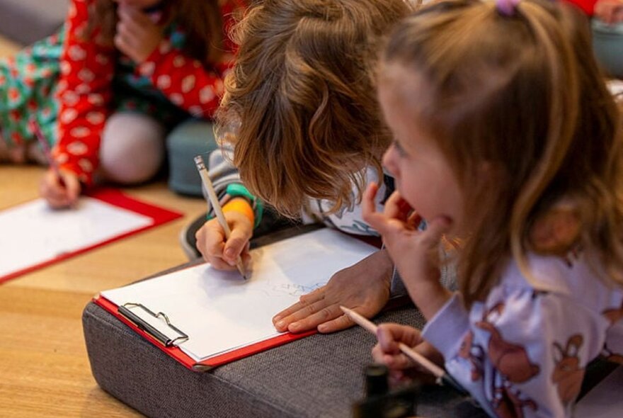 Three young children on the floor, each with a pencil and a clipboard.