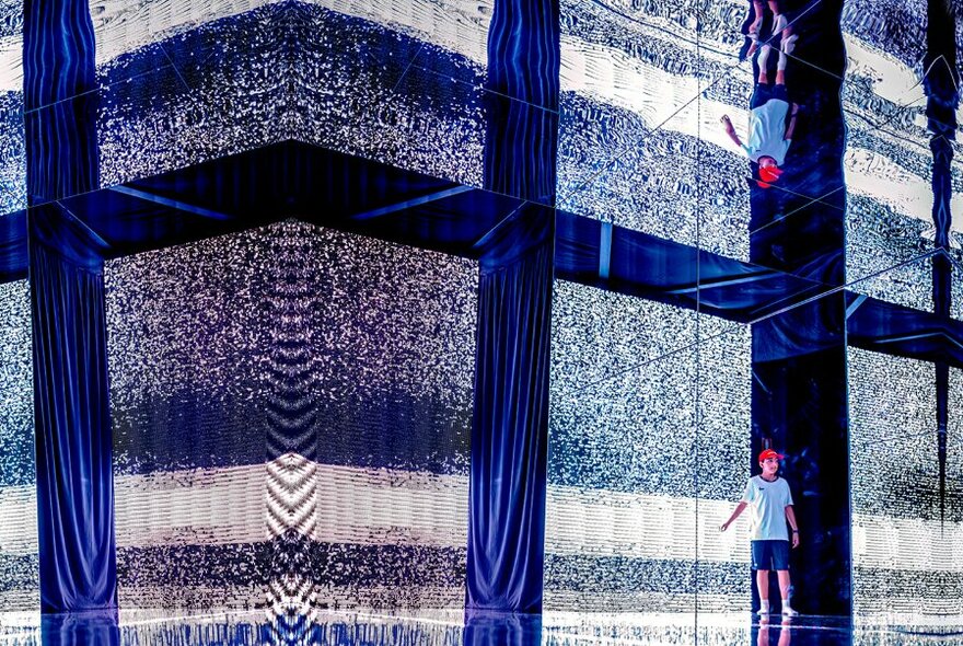 Person walking inside a space next to a digital display on a wall, a mirrored ceiling showing their reflection above. 