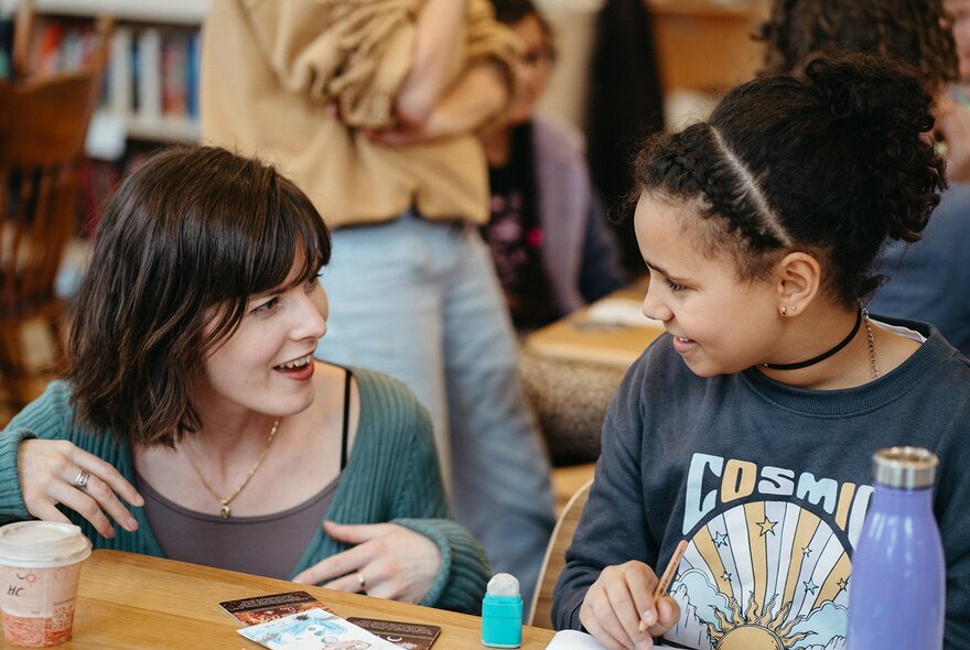An adult bobs down to table-height to speak with a girl working at a desk. 