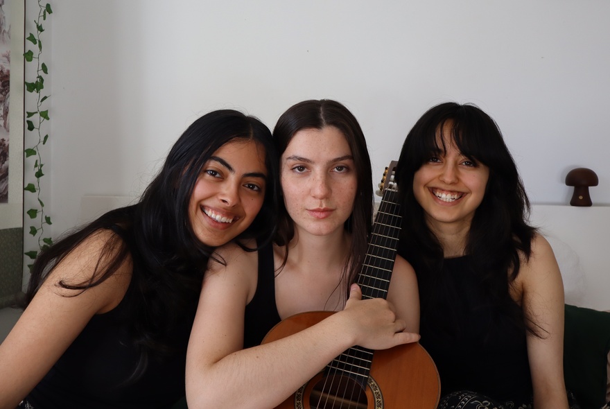 Three young women huddled together for a photo, the middle one holding a guitar and the other two smiling broadly. 