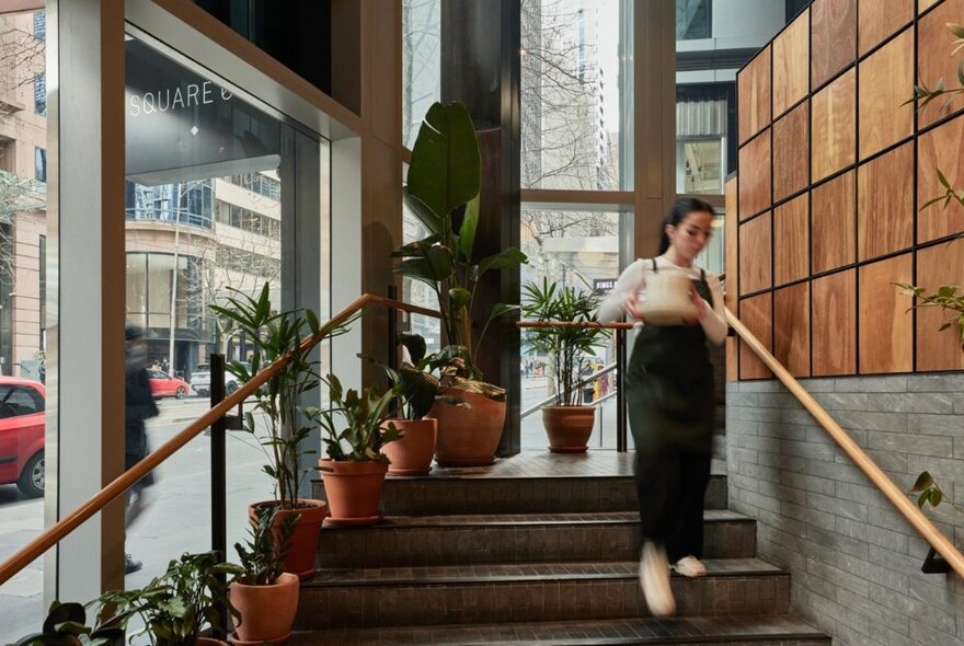 A person walking down stairs inside a cafe, carrying plates, large windows out onto the street to the left.