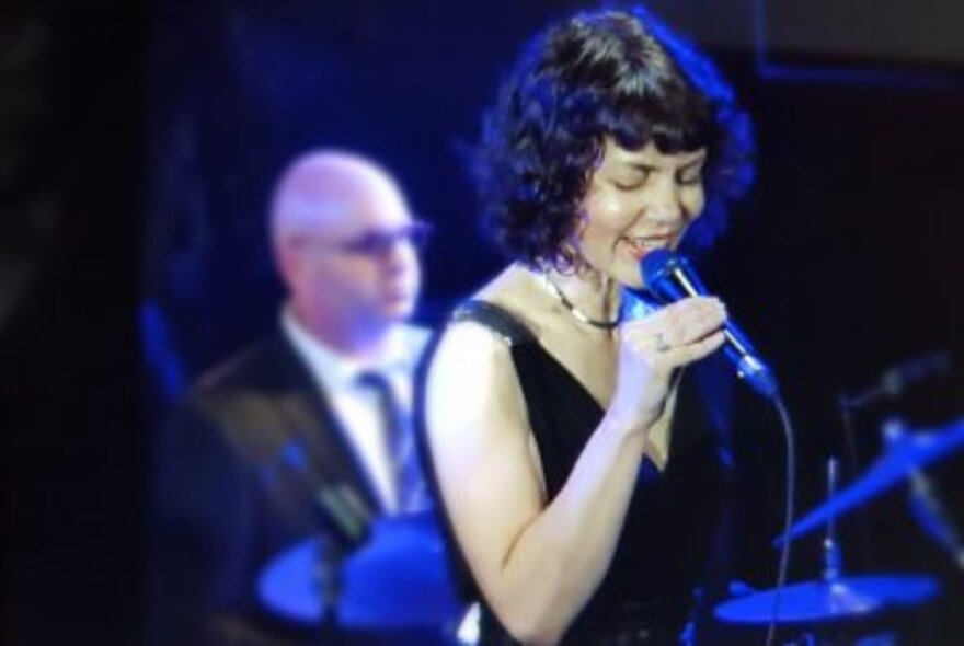 Woman with dark hair wearing a V-neck, sleeveless black dress performing on stage, singing into a microphone in front of the drummer.