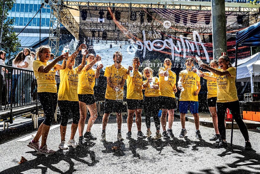 A group of 10 people in matching yellow t-shirts celebrating by shaking up their water bottles in the air in front of them, in an open air street setting.