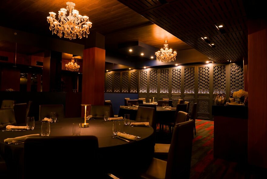 Moodily lit restaurant interior with dark-wood tables set for dinner beneath elaborate chandeliers.