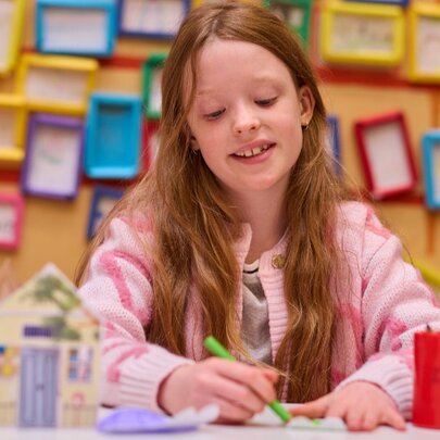A girl drawing with a green pencil in a crafting room with tens of coloured picture frames on the yellow wall behind her.