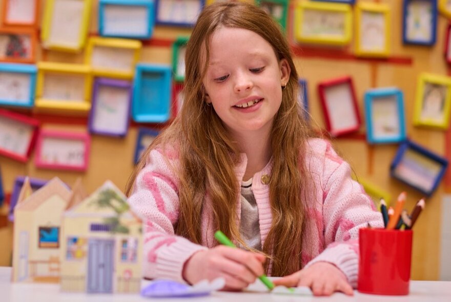 A girl drawing with a green pencil in a crafting room with tens of coloured picture frames on the yellow wall behind her.
